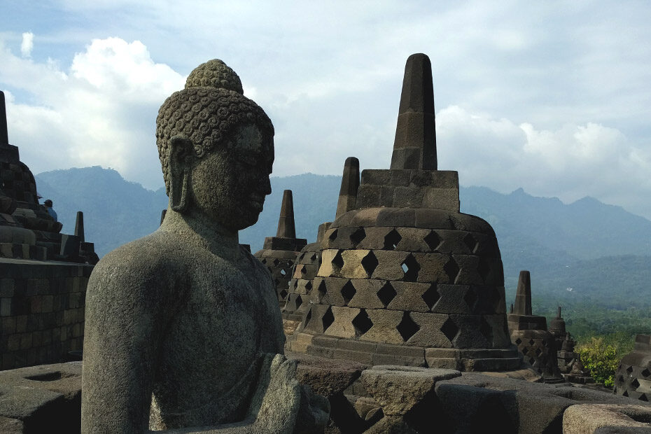 A state of the Buddha on the top level at Borobudur Temple in Indonesia. Stone stupas carved from volcanic rock are visible in the background. There are mountains visible in the distance and green jungle visible below at the bottom of the tiered temple.