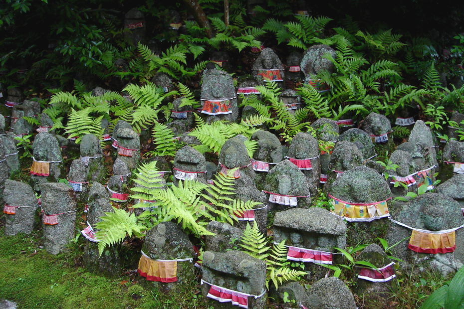 A cluster of old stone Jizo statues wearing red bibs, on a bed of moss, with bright green fern leaves poking out from amongst the stones.