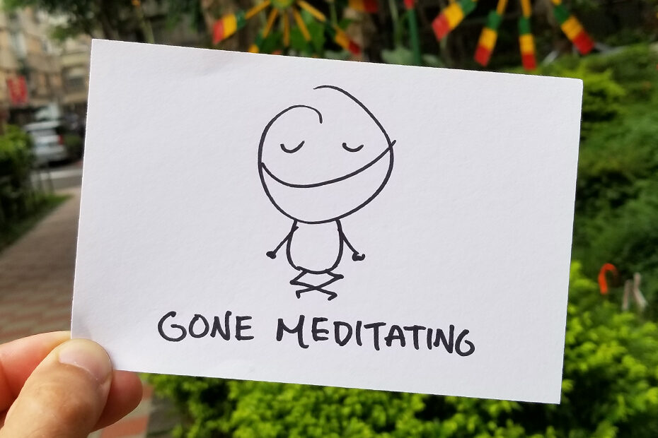 A drawing on a white piece of paper of a stick figure meditating with a big smile. Beneath them is written: GONE MEDITATING. Behind the note, trees and a street are blurry in the background.