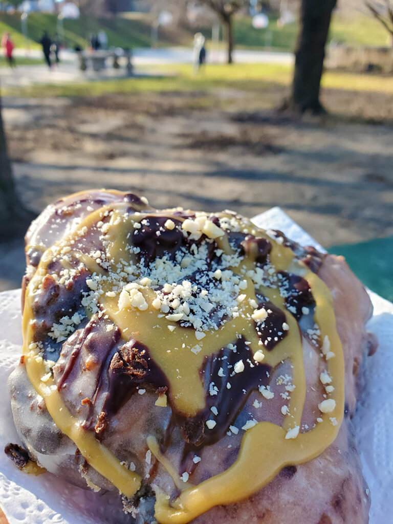 A Snickers fritter donut with caramel swirls, chocolate icing, and nuts sprinkled on top, with a park visible in the background