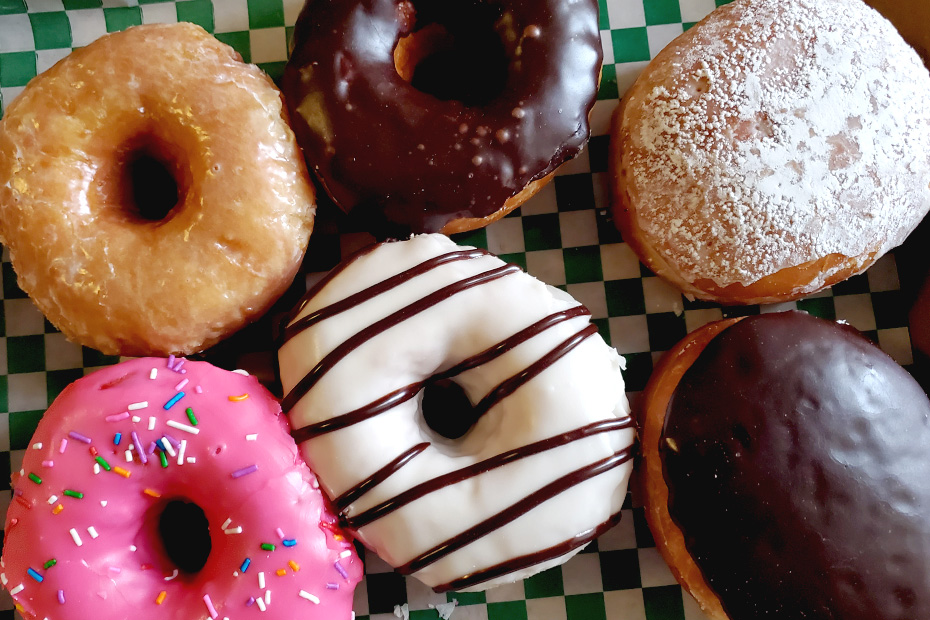 A variety of six of Toronto's best donuts in a box, with pink sprinkles, a yellow one, one covered in white icing with chocolate drizzles, a chocolate donut, a powdered jelly-stuffed, and a chocolate covered Boston cream