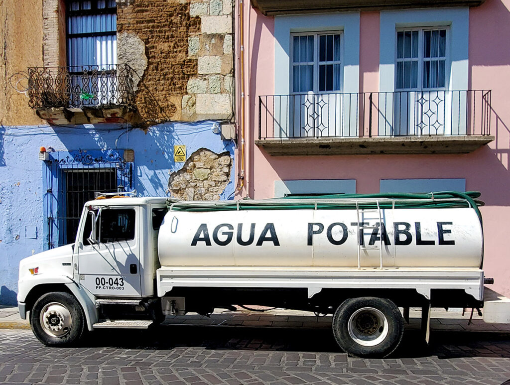 A tanker truck with AGUA POTABLE written on the side pumps drinking water into a home.