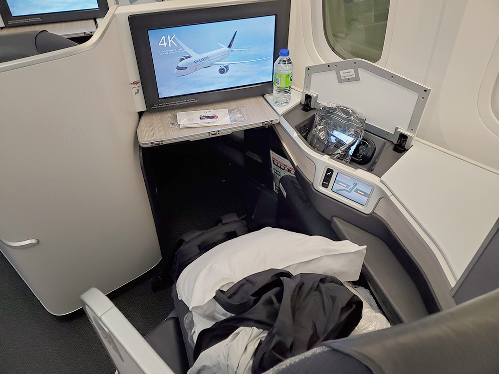 Air Canada Business Class seat 4K, with a screen displaying an image of the airplane, a compartment with headphones in plastic, a white pillow, Mark's rain coat on his seat, and a white blanket visible under that. The CleanCare+ sanitary kit is resting on a ledge in front of the entertainment system screen.