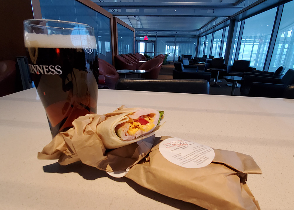 With a dark airport lounge behind it, on a table there is a Guinness glass full of beer and a ham and cheese wrap in a paper bag.