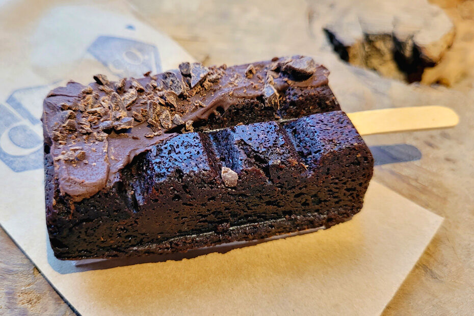 A brownie shaped like a paleta, with a popsicle stick jutting out from one end. It is a dark brown chocolatey color, with chocolate coating part of it, and crushed chocolate pieces captured in the drizzle. It is resting on a paper bag with the branding of Cho & Co.