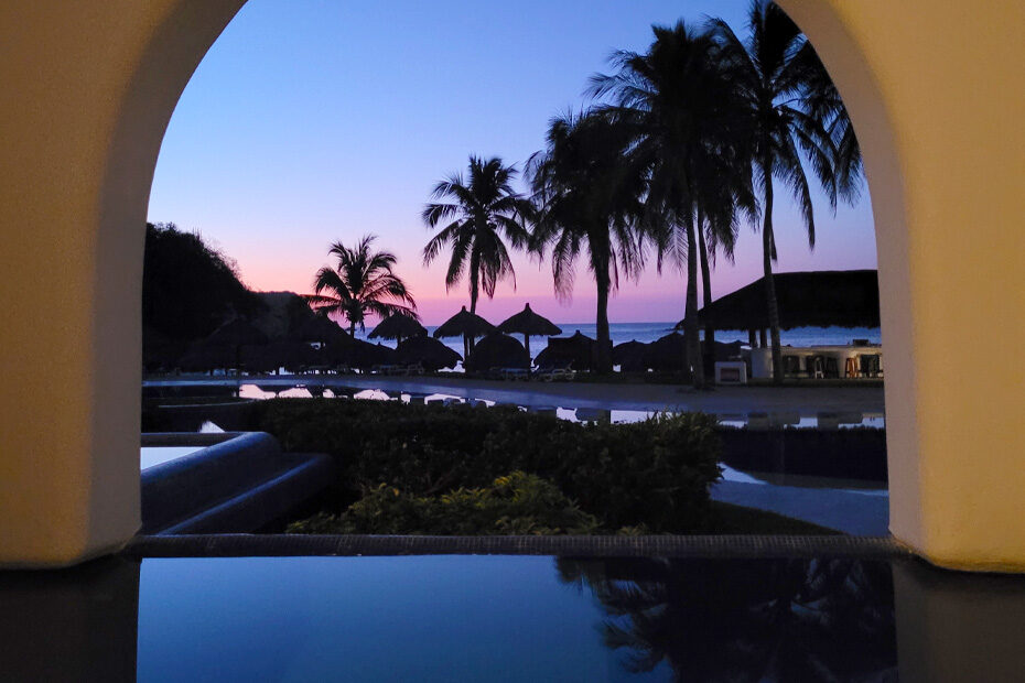 The pool at the Camino Real Zaashila, in the early morning. Palm trees are silhouetted against a pink and blue sky over the beach in the distance.