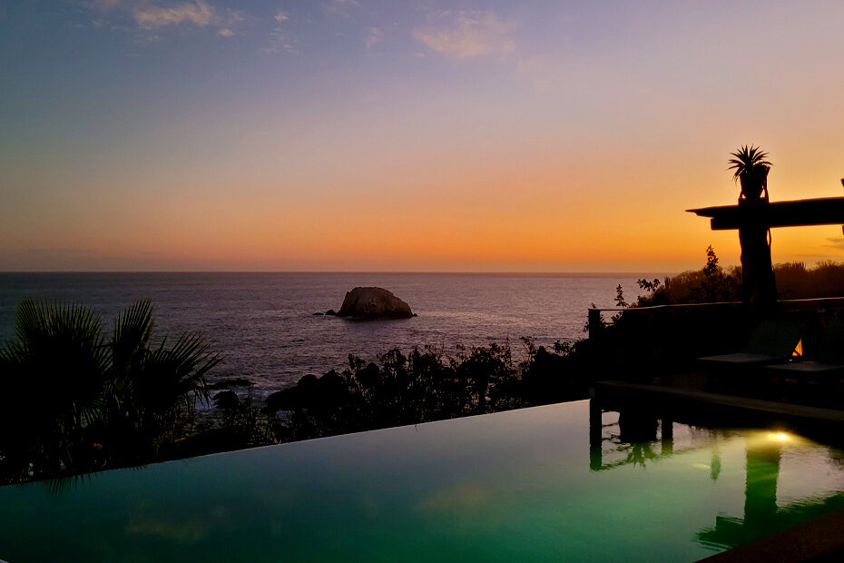 Sunset at Casa Kal-Mar, a bed-and-breakfast in Zipolite, Mexico. The sky is turned orange at the horizon. The infinity pool in the foreground is glowing blue and green. There are rocks in the ocean down below. Lounge chairs by the pool silhouetted against the sunset.