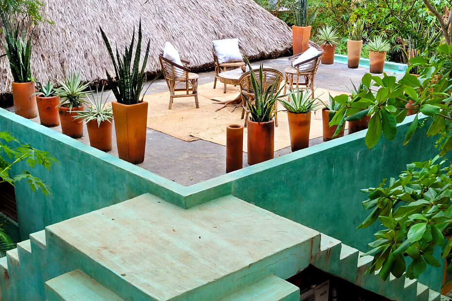 An open air, rooftop terrace at Monte Uzulu Hotel. There are four wicker chairs in the center with plush white pillows. The edges of the terrace are lined with terracotta pots of many different sizes, holding a variety of succulent plants. There are three staircases leading up to this terrace and they all meet at the corner on a square platform.