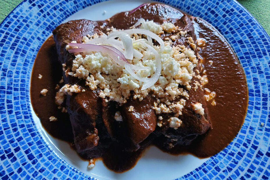 Enchiladas con Coloradito covered in a thick brown chocolate mole, garnished with crumbled cheese and onions.
