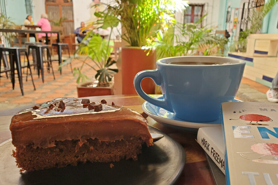 A piece of chocolate cake with thick icing on top, beside two books, and a mug of coffee as large as the cake, sitting on a table in the courtyard of my favorite coffee shop in Oaxaca, Mexico: Cafebre.