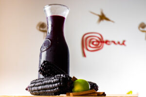 A glass carafe of deep purple chicha morada, with purple corn, limes, and cinnamon sitting on the table beside it, and Peru's tourism logo visible in the back. Copyright Shuttersstock