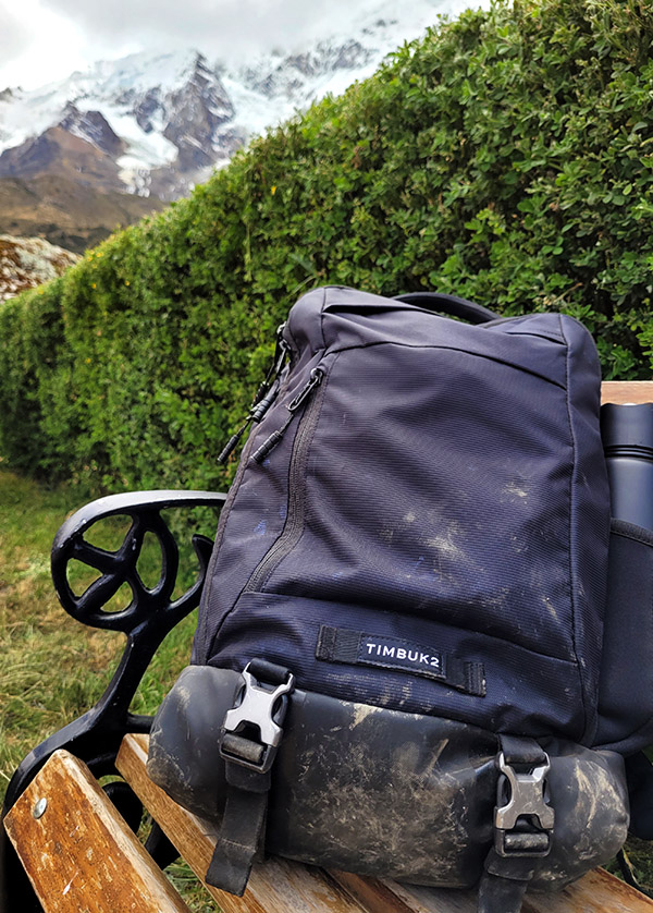 Timbuk2 Authority Laptop Backpack covered in mud, on a bench, with a green hedge behind it, and the snow-capped peaks of Humantay behind that