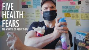 Image says: Five Health Fears and how to handle them. The words are beside a photo of Mark wearing a face mask, holding disinfectant spray in one arm that crosses his chest, pressing a container of wet wipes against him. His other arm is crossed under the disinfectant spray, holding a squeeze bottle of hand sanitizer, finger ready to press down on the bottle's dispenser. His eyes look concerned, one eyebrow raised.