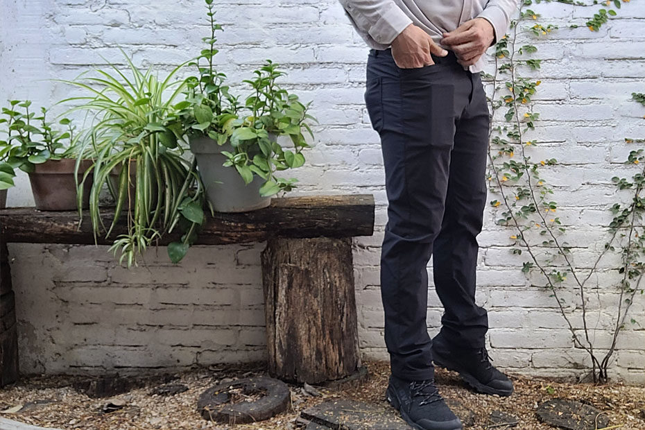 Wearing the Western Rise Evolution Chino, black, over Inov8 Roclite boots, standing outside against a brick wall painted white, with plants in pots on a wood bench